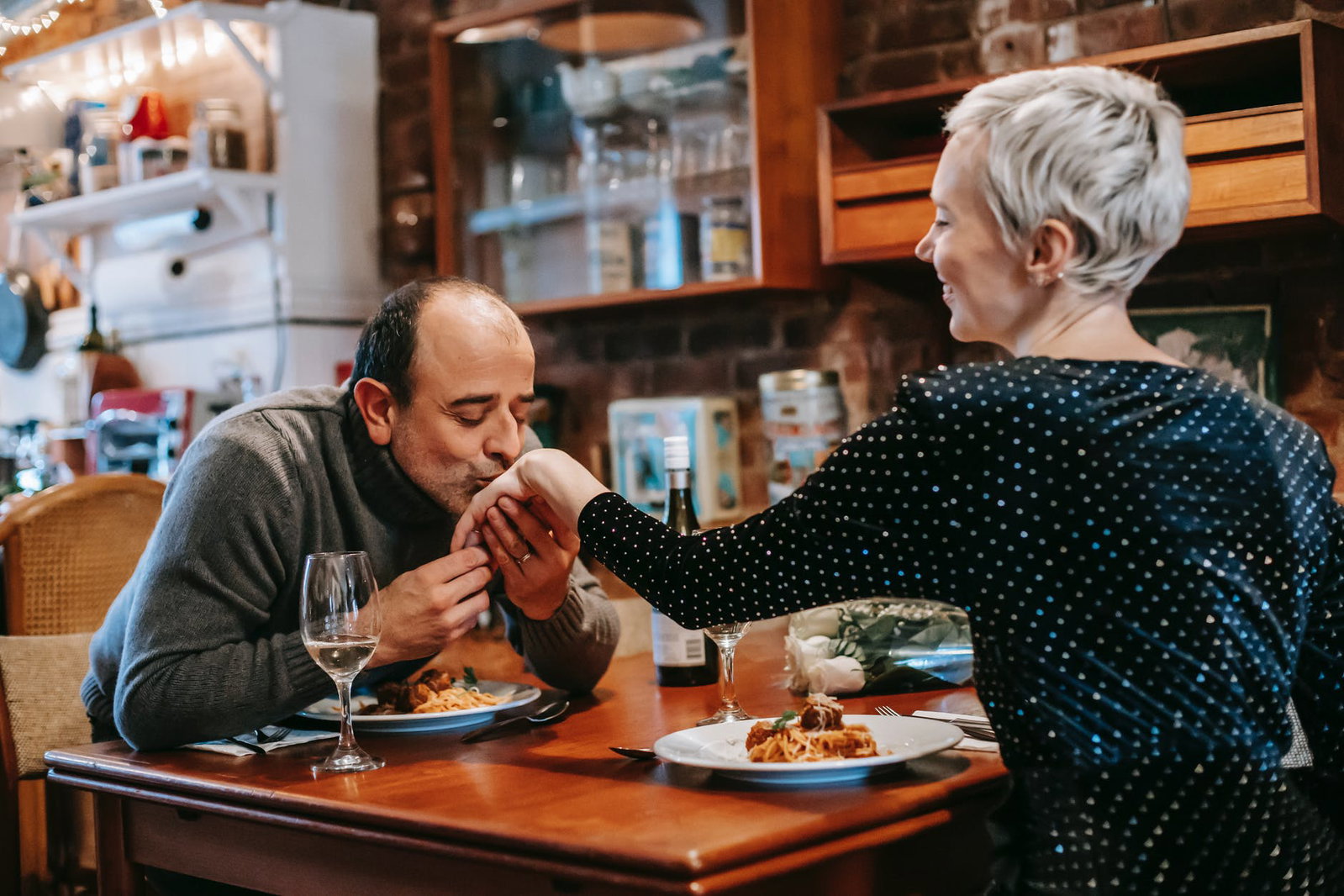 man and woman sitting at a dining table with food as he kissed her hand