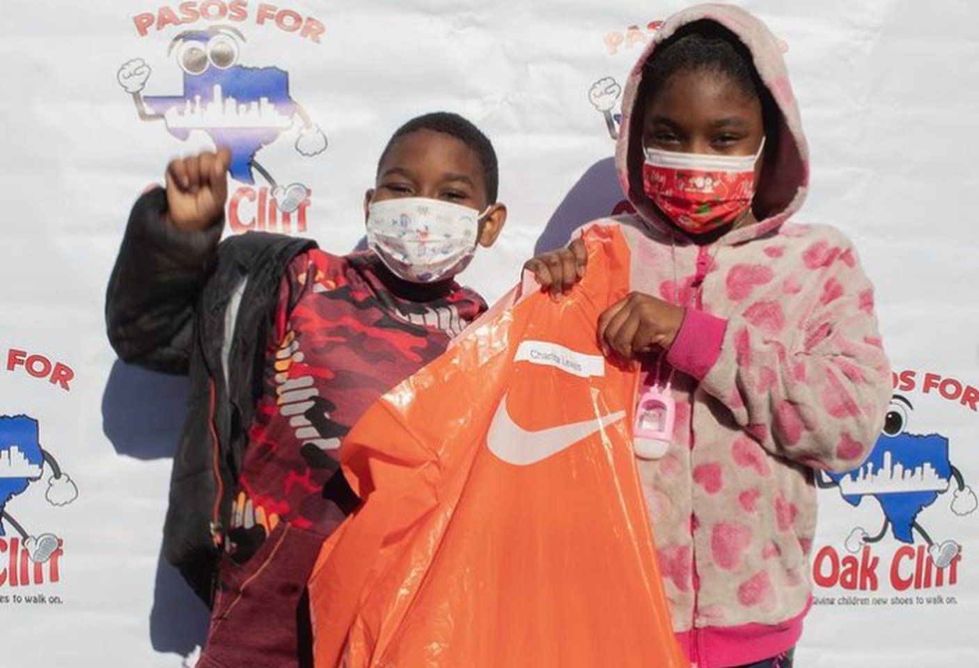 little boy and little girl wearing masks as they excitedly hold an orange nike bag with new sneakers at a pasos for oak cliff shoe drive