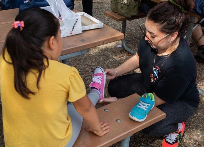 little girl sitting on a bench outside as a woman helps her try on a pink pair of sneakers at a pasos for oak cliff shoe drive
