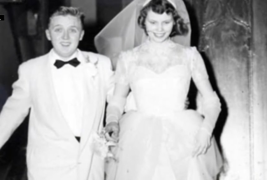 black and white photo of kenneth and elizabeth gage smiling as they walk together in their wedding tux and dress. 