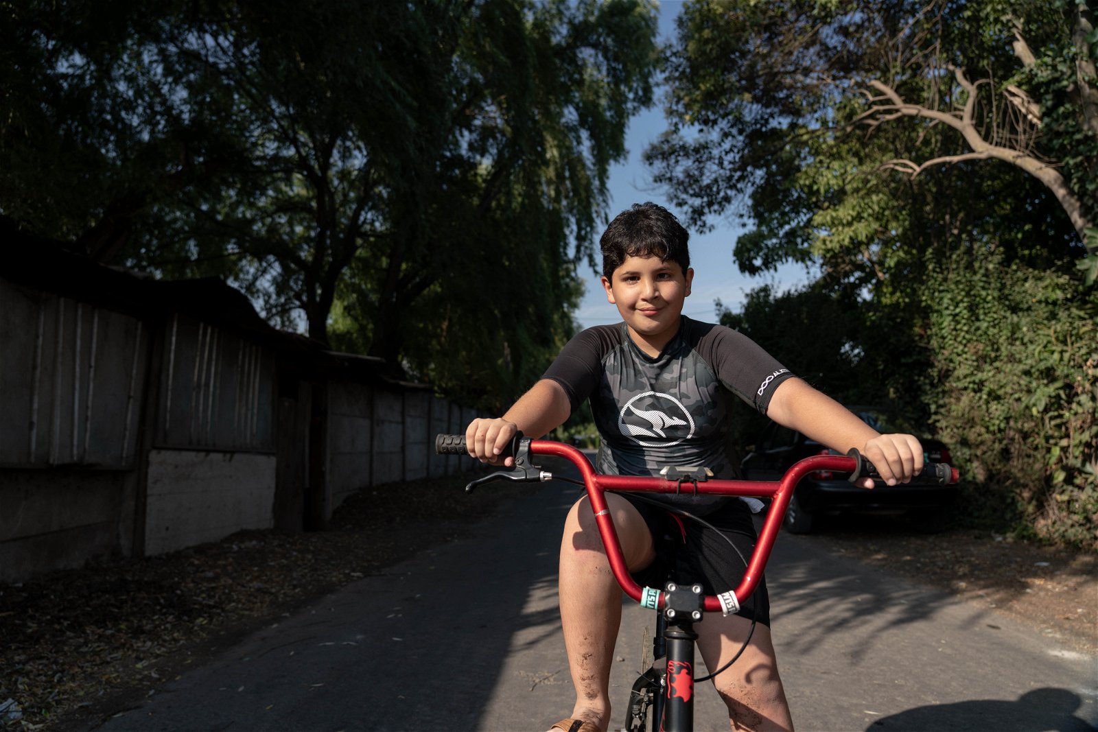 juan smiling as he rides his red bike down a road with trees on each side.