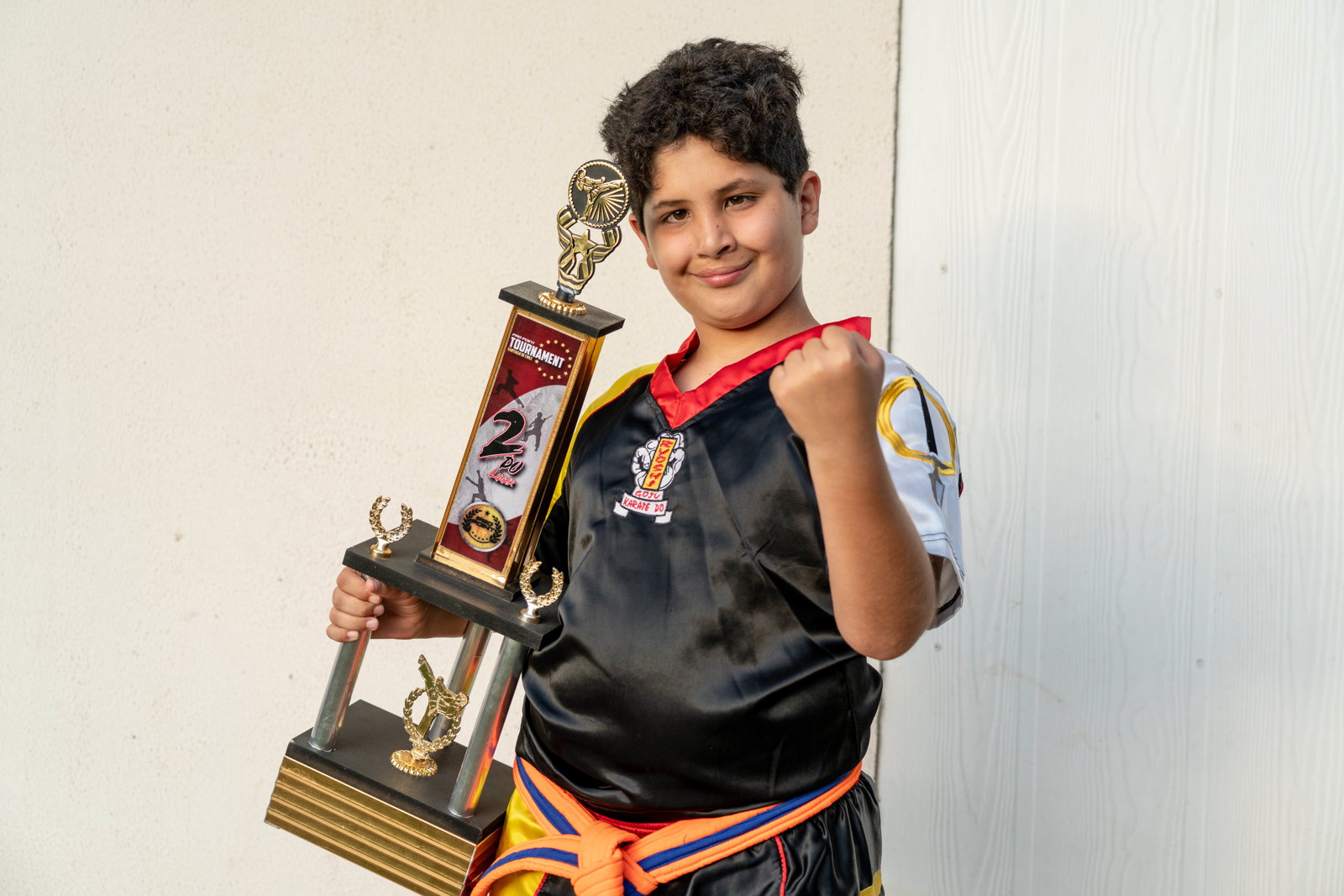 juan smiling. he's posing with his large taekwondo trophy in one hand while, with the other, he does a taekwondo pose. he's wearing his taekwondo uniform.