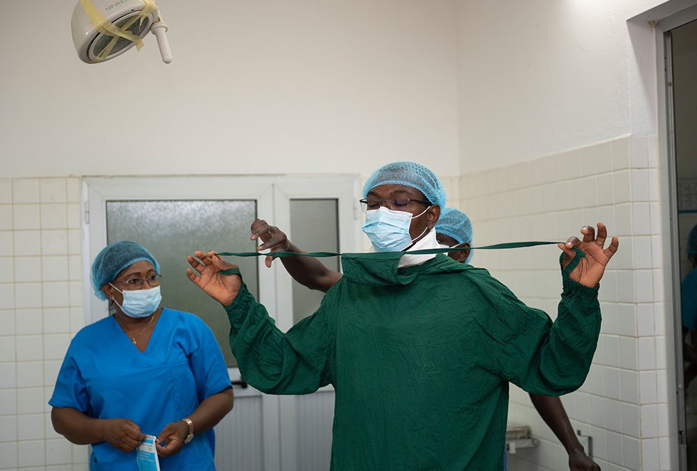Dr. GrÃ©goire Akakpo-Numado being assisted while putting on scrubs before cleft surgery.