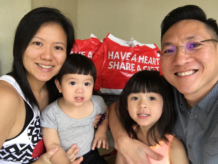 Francis, Jolene, and two of their kids smiling as they pose for the photo. Behind them are red and white bags that read, "Have a heart, share a gift." They come from The Boys’ Brigade Share-a-Gift Project.